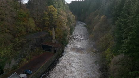 Drone-footage-flies-high-above-a-fast-flowing-river-before-descending-towards-a-waterfall-surrounded-by-buildings-and-a-forest-of-broadleaf-and-coniferous-trees