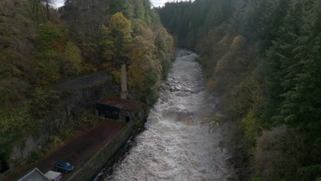 imágenes de drones vuelan alto sobre un río que fluye rápido antes de descender hacia una cascada rodeada de edificios y un bosque de árboles coníferos y de hoja ancha