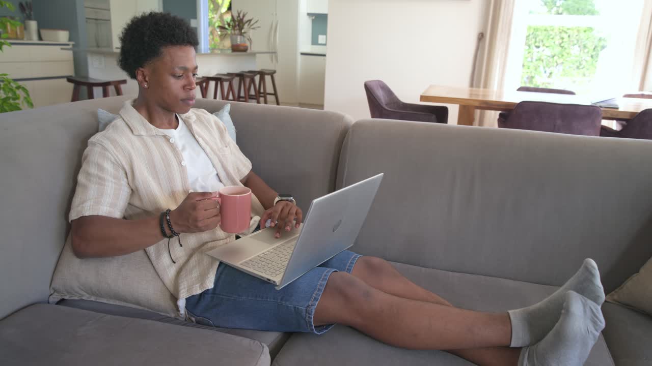 African American adult male working on laptop, sipping pink mug, typing, glancing at window