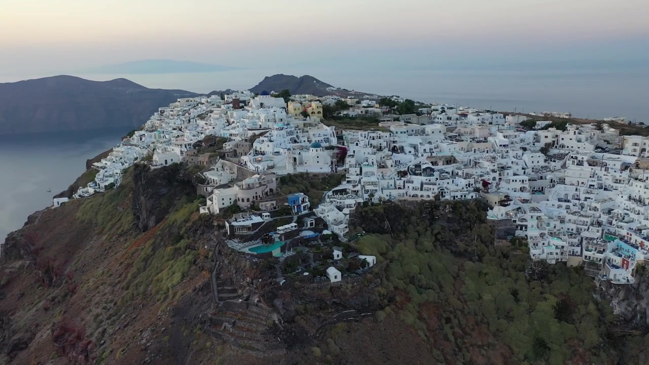 Impressionante tomada ampla de drone de Fira, Santorini, ao entardecer. Apresenta vilarejos brancos tradicionais construídos ao longo de enormes falésias vulcânicas com vista para iates de luxo ancorados no calmo Mar Mediterrâneo.