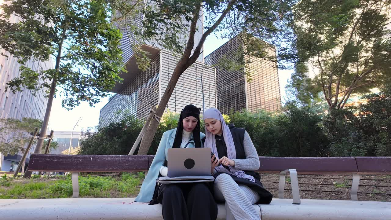 Two women using a laptop on a bench in the city