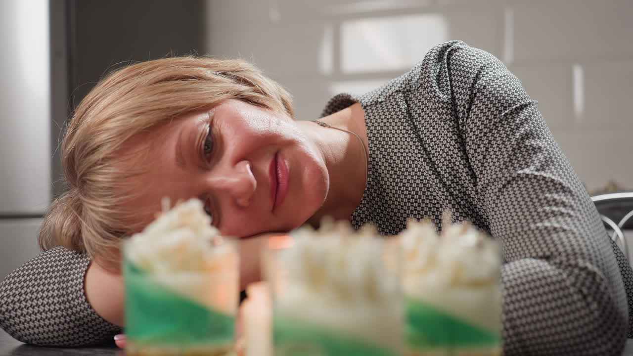 Elegant woman in patterned dress rests head on table with soft smile as she peacefully admires beautiful handmade candle jars, expressing warmth, satisfaction, and calmness in cozy kitchen atmosphere