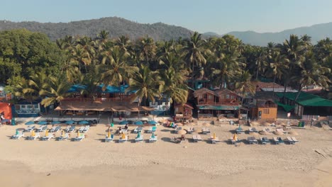 palolem beach resort shacks and resting chairs near the coastline in exotic setting - aerial fly backwards reveal