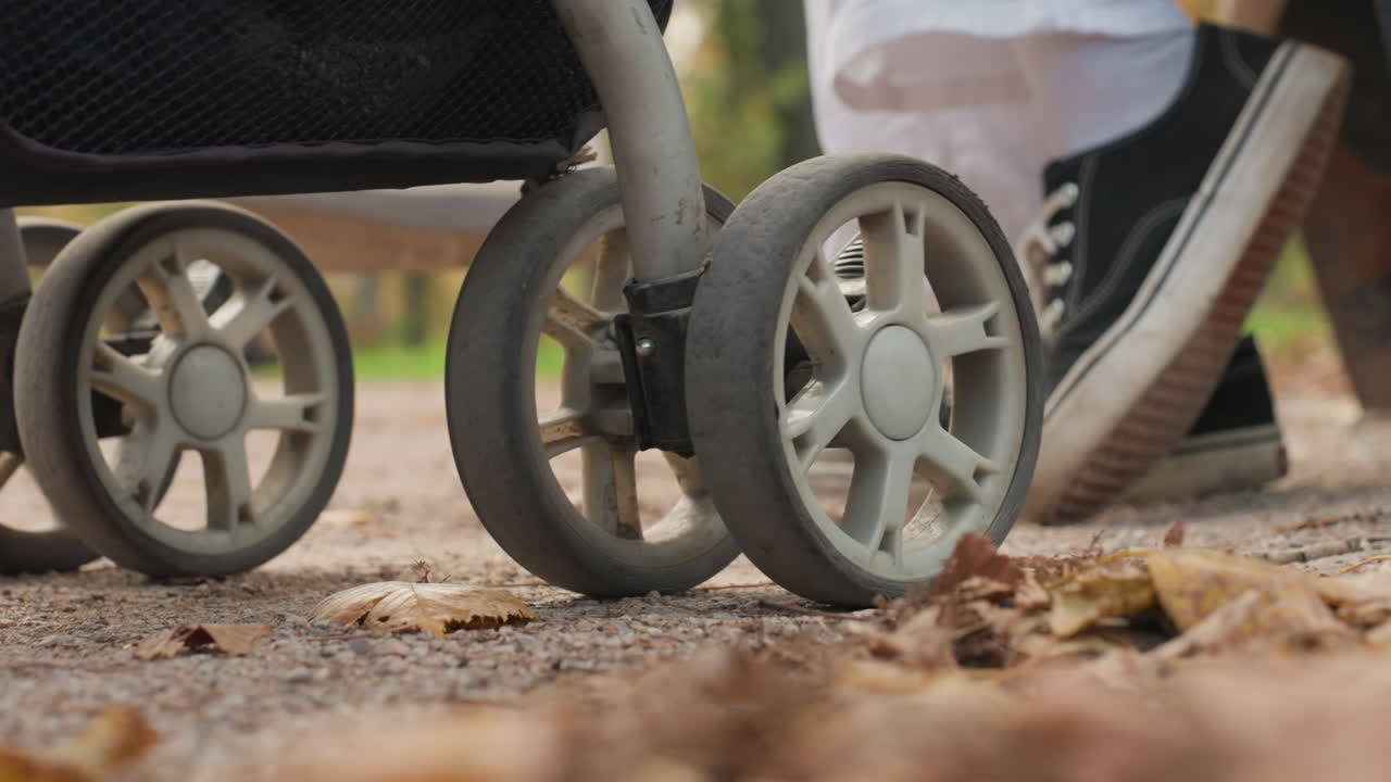 Närbild på barnvagnshjul på en stig. En förälder i sneakers kliver över fallna löv. Höstljus, en uppriktig slow motion-fångst, ett intimt familjeögonblick och ett tyst, varmt band som antyder äktenskap och föräldraskap.