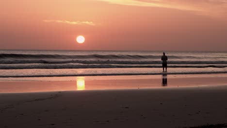 Roter-Sonnenuntergang-Am-Strand-Von-Playa-Maderas-In-Nicaragua-Mit-Einem-Mann,-Der-Steine-Auf-Die-Brandung-Wirft,-Weitwinkelaufnahme-Aus-Der-Hand