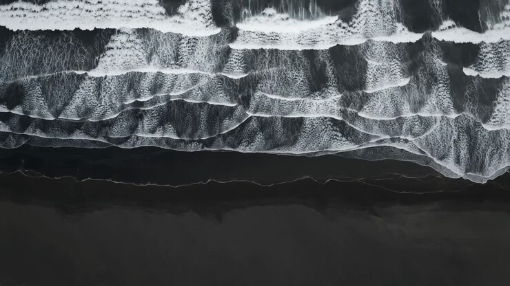 Vista aerea di onde oceaniche che si infrangono su una spiaggia scura, catturata dall'alto.