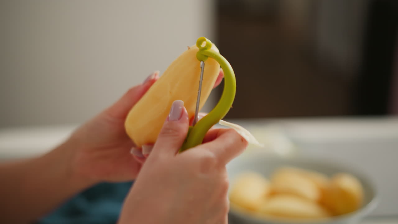 Close up of fair skinned person peeling potato with peeler, hands in focus, blurred plate of peeled potatoes in background on white kitchen counter, soft lighting
