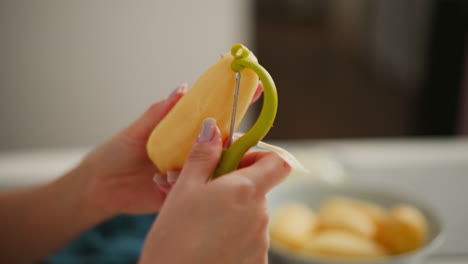 close up of fair skinned person peeling potato with peeler, hands in focus, blurred plate of peeled potatoes in background on white kitchen counter, soft lighting