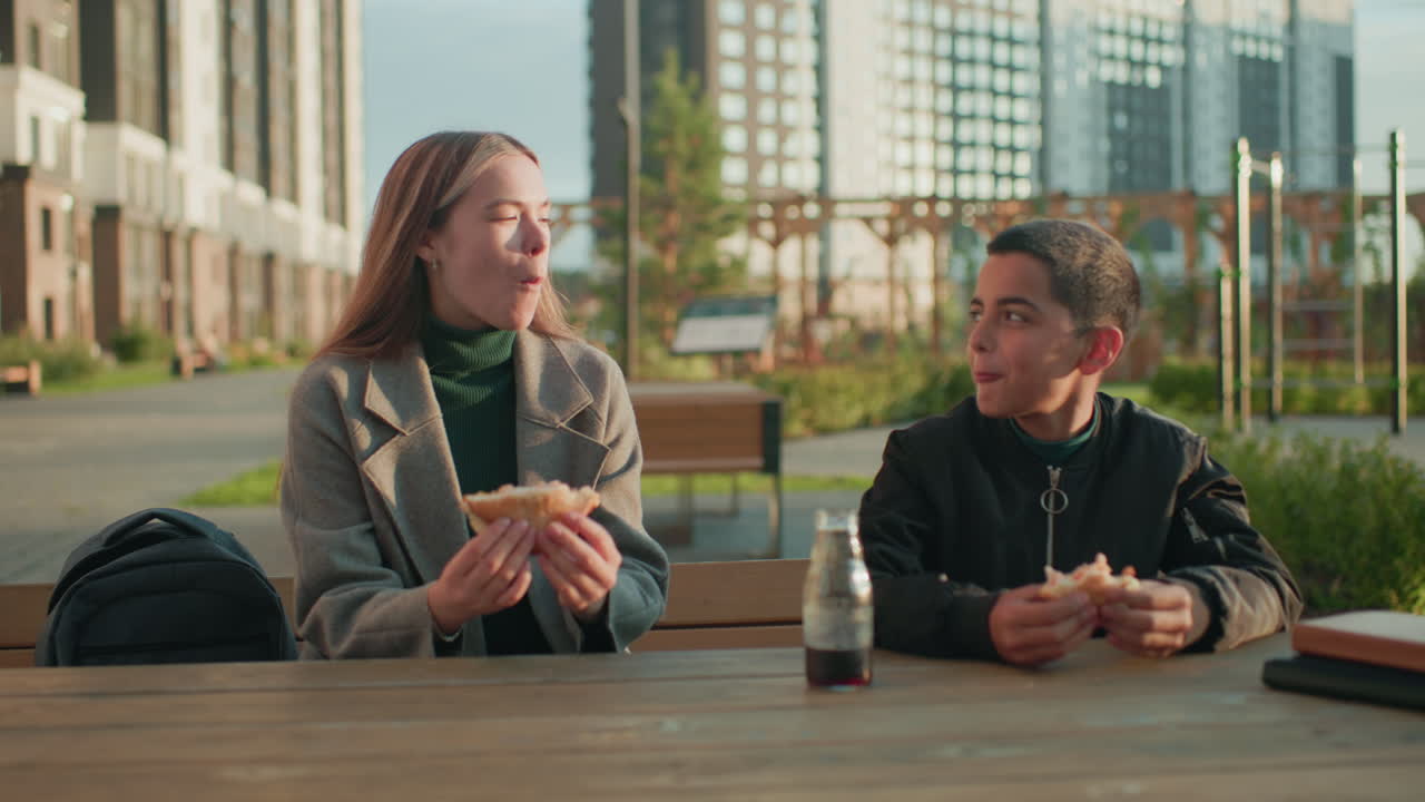 Lady and kid sitting outdoors at wooden table in modern urban park making toast with bread in hand while enjoying meal together, smiling warmly and bonding during snack time