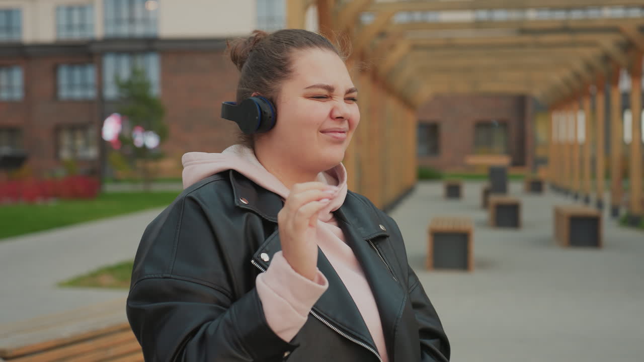 Lady puts on headphones and begins dancing with joy while making expressive facial gestures, enjoying music outdoors in urban park setting with stylish wooden structures and autumn ambiance