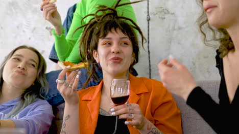 mujer feliz con tatuajes y rastas comiendo pizza y bebiendo vino mientras habla con amigos en casa 1