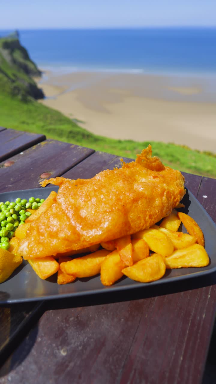 Vertical Video Classic British Fish and Chips on Wooden Picnic Table with Sea Beach Landscape Background on Clear Sunny Day. Illustration of Fresh Seafood from Boat to Plate. Served with Chips, Peas.