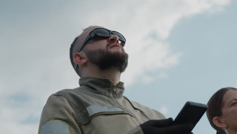 close up adult wearing dark gloves holding smartphone while companion gazes skyward with thoughtful expression against blurred green field backdrop under bright cloudy sky