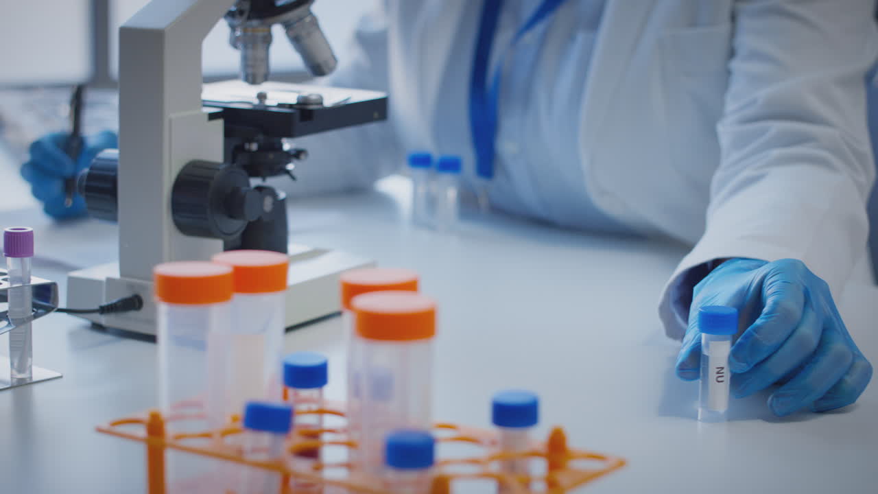 primer plano de un trabajador de laboratorio que lleva a cabo investigaciones utilizando un microscopio sosteniendo un tubo de ensayo etiquetado nu