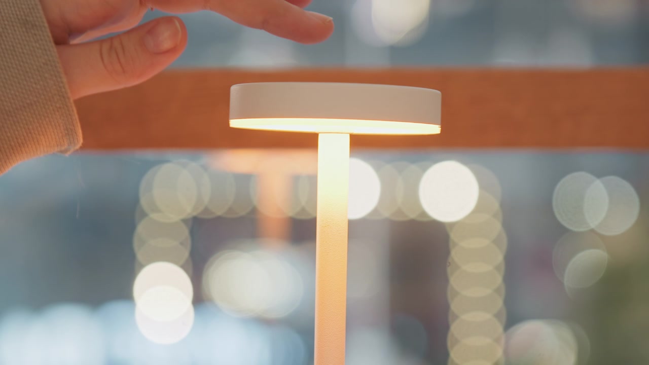Nærbilde av en hånd som slår på en elegant hvit LED-lampe som avgir mykt lys i et moderne innendørsmiljø. Scenen har varme omgivelseslyseffekter fra en uskarp bakgrunn.