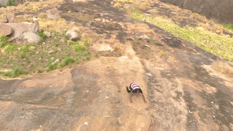 Toma-Aérea-Mirando-Hacia-Un-Acantilado-De-Granito-Mientras-Un-Joven-Africano-Sube-Por-La-Cara-Escarpada