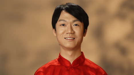 카메라를 보고 웃는 붉은 전통 의상을 입은 쾌활한 젊은 아시아 남자의 클로즈업 보기