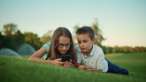 children using smartphone in park. siblings lying on green grass in meadow