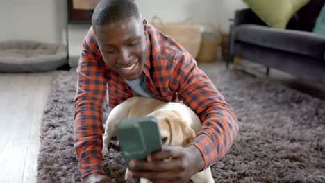 Happy-african-american-man-lying-on-floor-using-smartphone-at-home,-with-his-pet-dog,-slow-motion