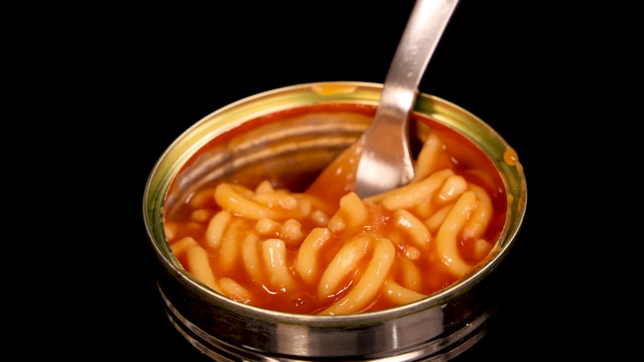 A metal fork lifts spaghetti in tomato sauce from an open can, isolated on a black background with even, bright lighting and close-up framing
