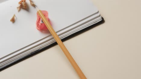 Close-up-of-open-notebook-with-pencil-and-pencil-sharpener-on-beige-background,-in-slow-motion