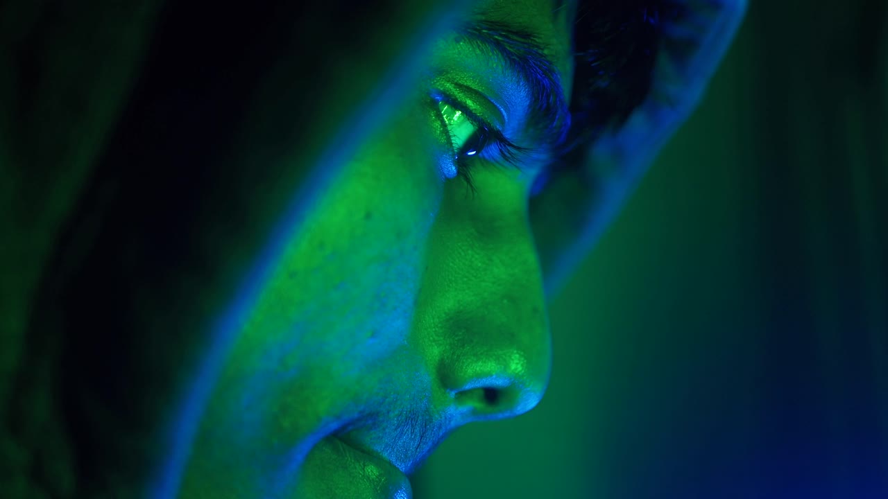 Close-up portrait of a person wearing a hoodie, illuminated with green and blue light.
