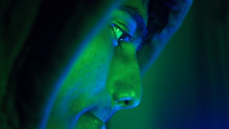 Close-up portrait of a person wearing a hoodie, illuminated with green and blue light.