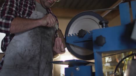 hombre caucásico fabricante de cuchillos en un taller con gafas y usando una lijadora