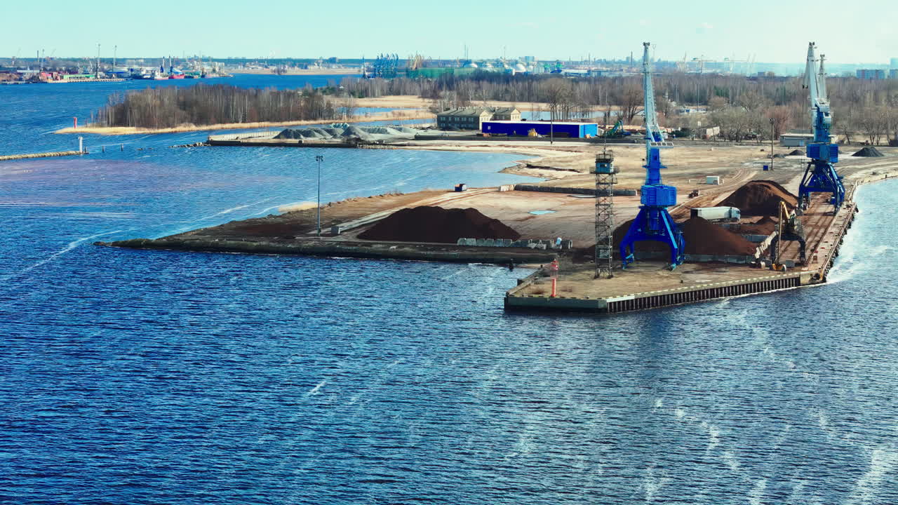 A drone glides above an industrial harbor where blue cranes service bulk cargo stockpiles on a narrow pier, choppy water and distant shoreline framing a busy, weathered waterfront scene