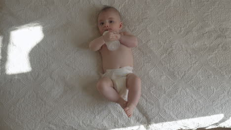 Newborn-baby-holding-his-milk-bottle-independently,-laying-on-back-on-white-bed-sheets