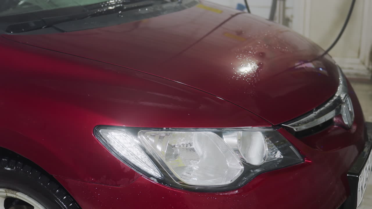 close up of red car bonnet being rinsed in garage with bright light reflection highlighting glossy paint surface and water droplets with visible steam enhancing metallic sheen on body