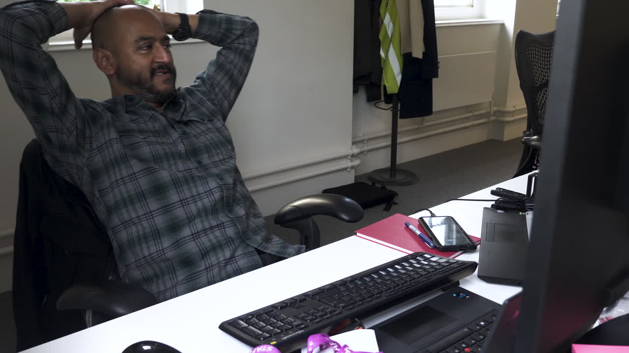 A close up shot of an Asian Indian male relaxed leaning back on an office chair with his hands on his head while casually talking to colleagues in the room