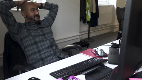 a close up shot of an asian indian male relaxed leaning back on an office chair with his hands on his head while casually talking to colleagues in the room