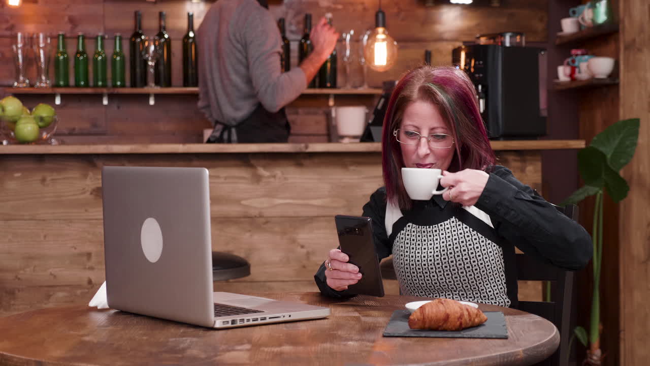 Woman Using Smartphone and Laptop in Cafe with Coffee and Croissant