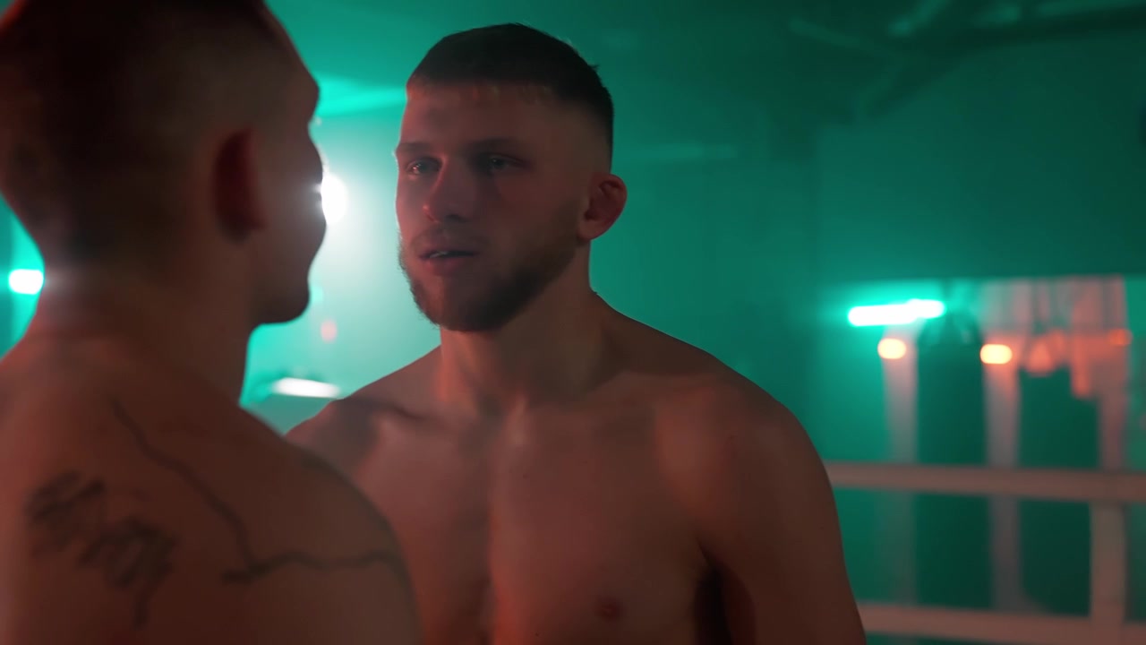 Due pugili a torso nudo che si fissano intensamente in un ring da boxe buio.