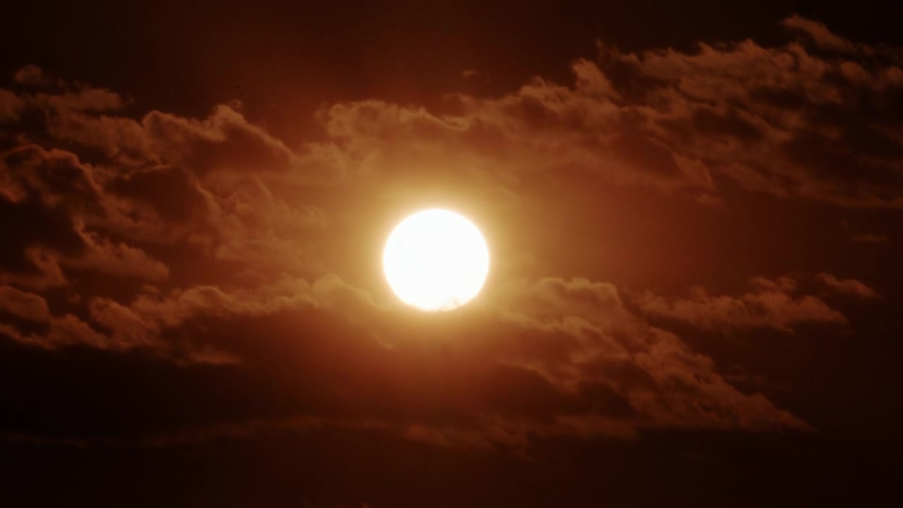 Närbild av en stark sol delvis täckt av tjocka rörliga moln, som glöder i intensiva guldorange solnedgångstoner.