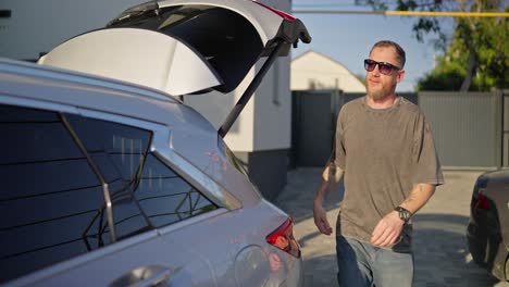 Confident-guy-in-sunglasses-with-his-girlfriend-putting-bags-in-the-trunk-of-the-car-and-getting-into-the-cabin-while-leaving-the-hotel-in-summer