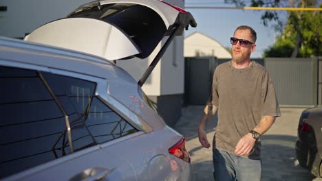 hombre confiado en gafas de sol con su novia poniendo bolsas en el maletero del coche y entrando en la cabina mientras sale del hotel en verano