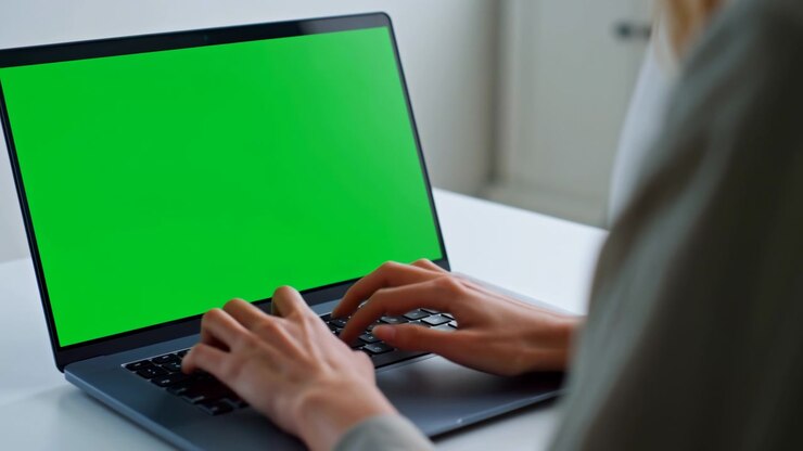 Le dita si muovono sui tasti neri mentre il display verde fisso del portatile rimane invariato, incorniciato dal case grigio, dal tavolo bianco e da una spalla sfocata in un ambiente interno leggermente illuminato