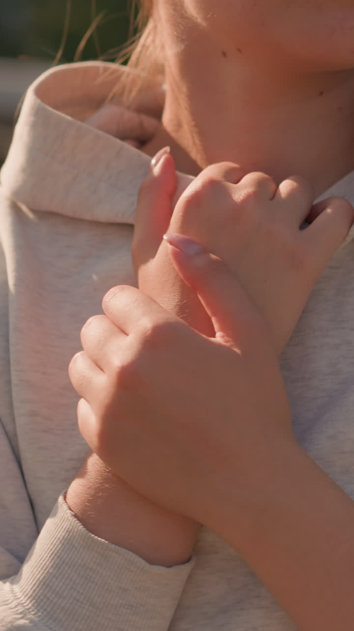 Close-up of woman crossing her hands over her hoodie collar, gently touching her neck in a reflective, serene moment, sunlight softly illuminates her skin