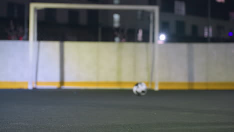 vista traseira de um homem habilmente chutando uma bola de futebol para o poste de gol com precisão durante a sessão de prática noturna, fundo apresenta cerca metálica, edifício residencial iluminado