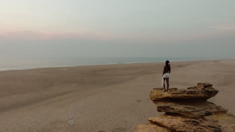 aerial shot orbiting a woman standing barefoot on a coastal rock, facing the hazy horizon at dusk