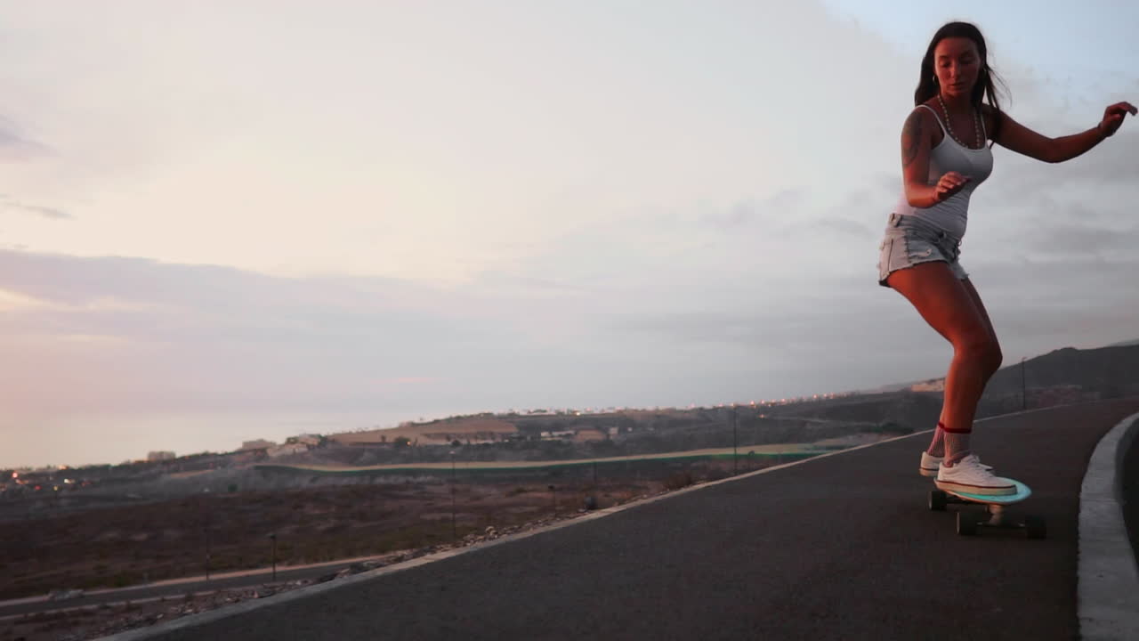 A mountain road provides the setting for a stylish and beautiful skateboarder to ride her board in shorts during sunset, with the mountains' incredible view showcased in slow motion