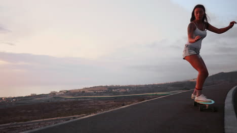 a mountain road provides the setting for a stylish and beautiful skateboarder to ride her board in shorts during sunset, with the mountains' incredible view showcased in slow motion