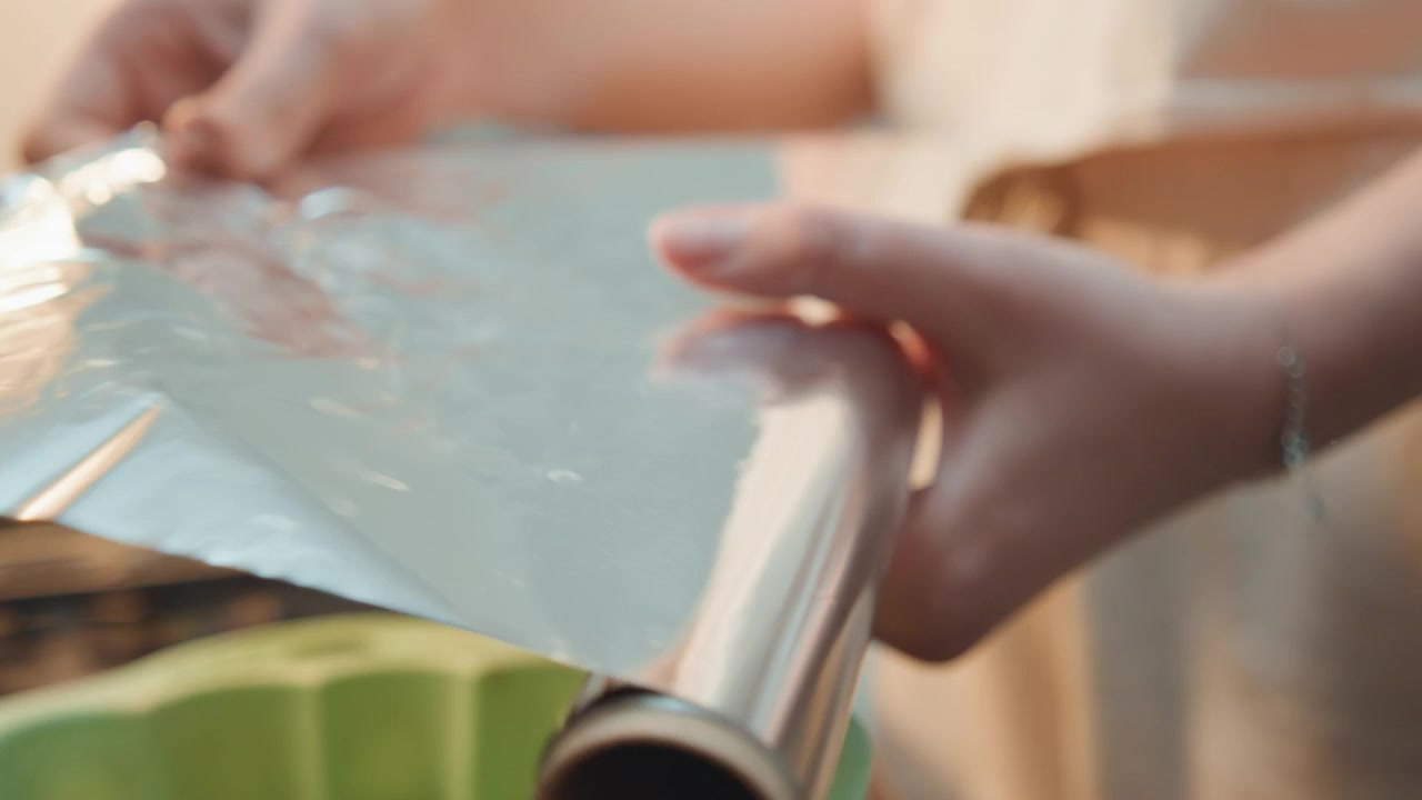 Mani che srotolano la carta stagnola su una teglia verde sul fornello, primo piano del rotolo lucido e dei bordi netti, preparazione accurata per la cottura, movimento ordinato del polso e tiro misurato, stampo in silicone per ciambella pronto per l'impasto.
