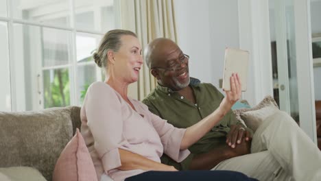 Happy-senior-diverse-couple-wearing-shirts-and-using-tablet-in-living-room