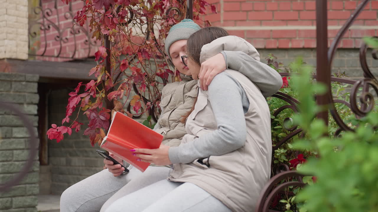 Side view of siblings seated outdoors in cool breeze as man warmly brings sister close while she reads book and holds phone, surrounded by lush garden, red autumn leaves, and decorative metal bench