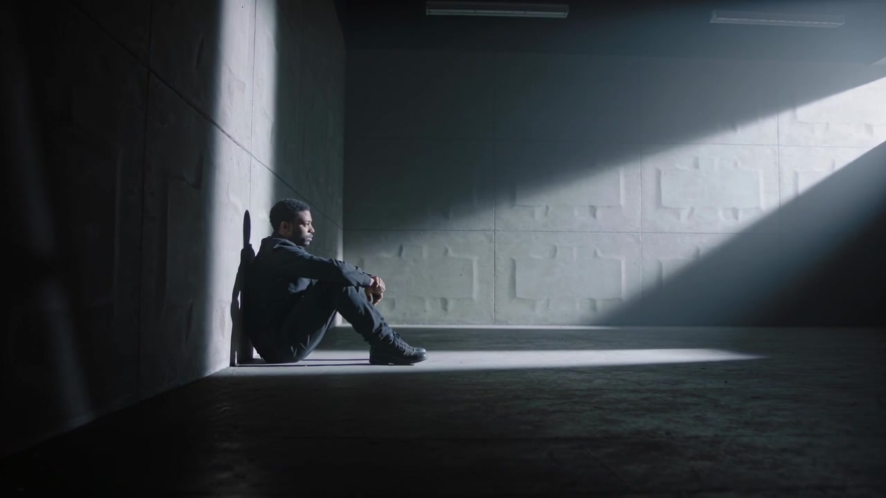 Turning toward light shaft, man waiting in concrete room in dark hoodie rifle beside, copy space