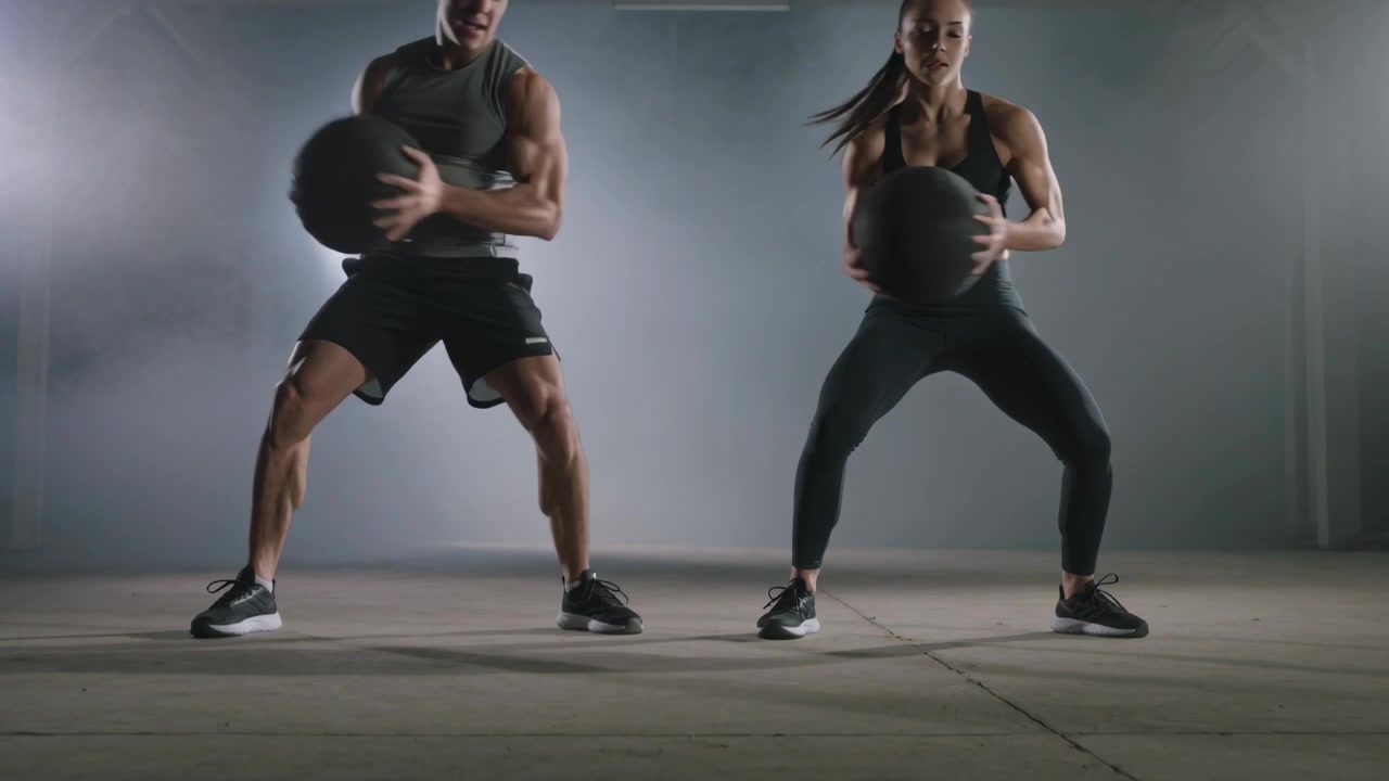 Uomo a sinistra e donna a destra eseguono una routine speculare con la palla medica in palestra, allenandosi in abbigliamento sportivo.