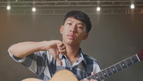 primer plano de un adolescente asiático con una guitarra mostrando el pulgar hacia abajo mientras está de pie en el escenario